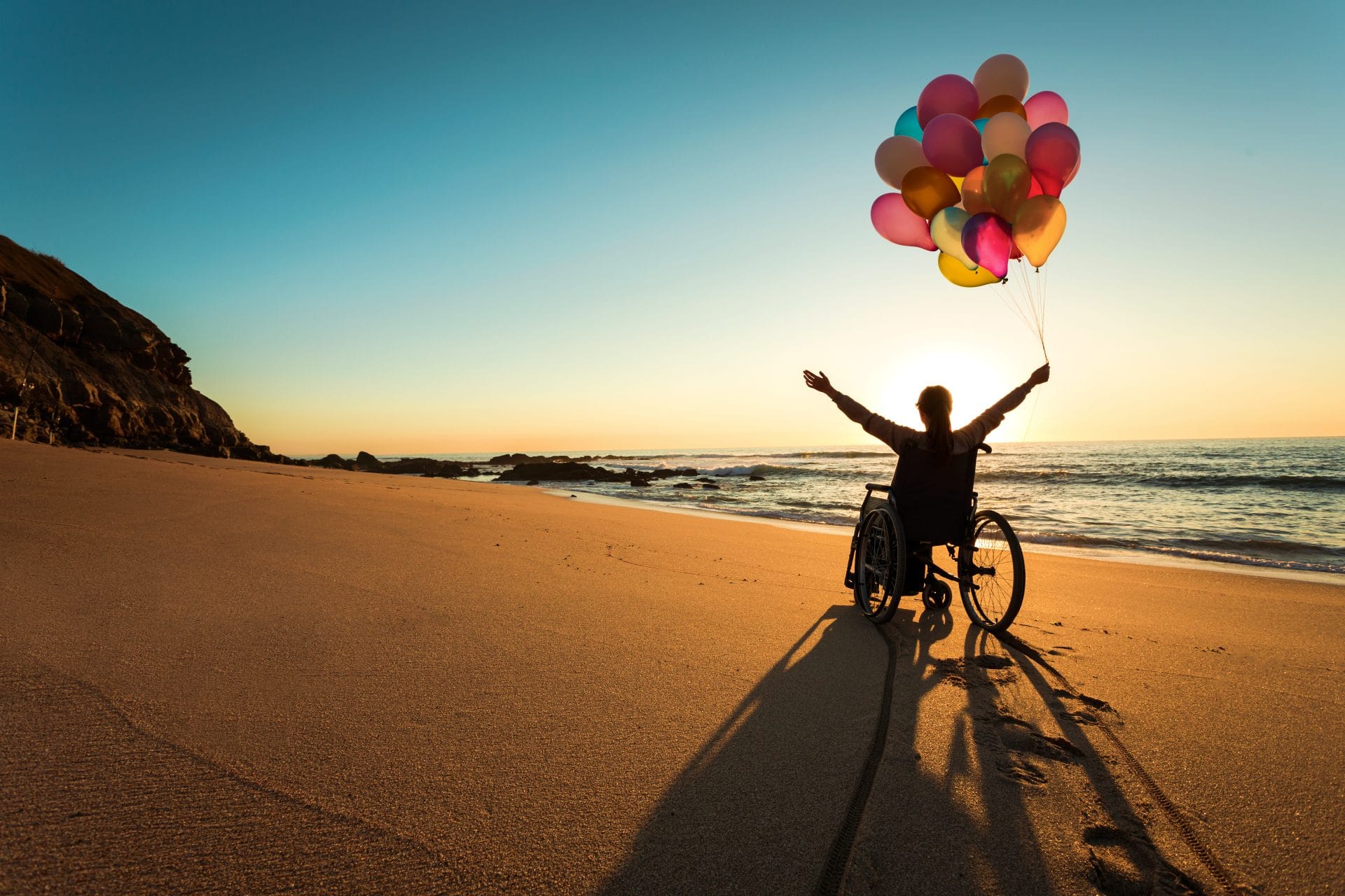 A woman using a wheelchair raises her arms with balloons on a beach at sunrise